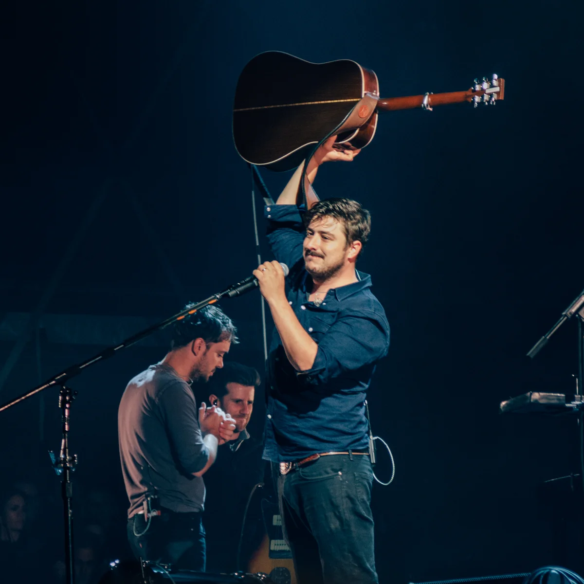 Marcus Mumford holding a guitar above his head during a public performance in Aviemore in 2015